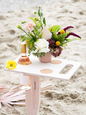 Goulburn portable beach table in beach blonde color unfolded standing up in sand at beach with refreshment drinks and floral decor