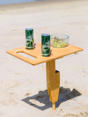Goulburn portable beach table in naturally sunkissed color unfolded standing up in sand at beach with refreshment drinks and fruit