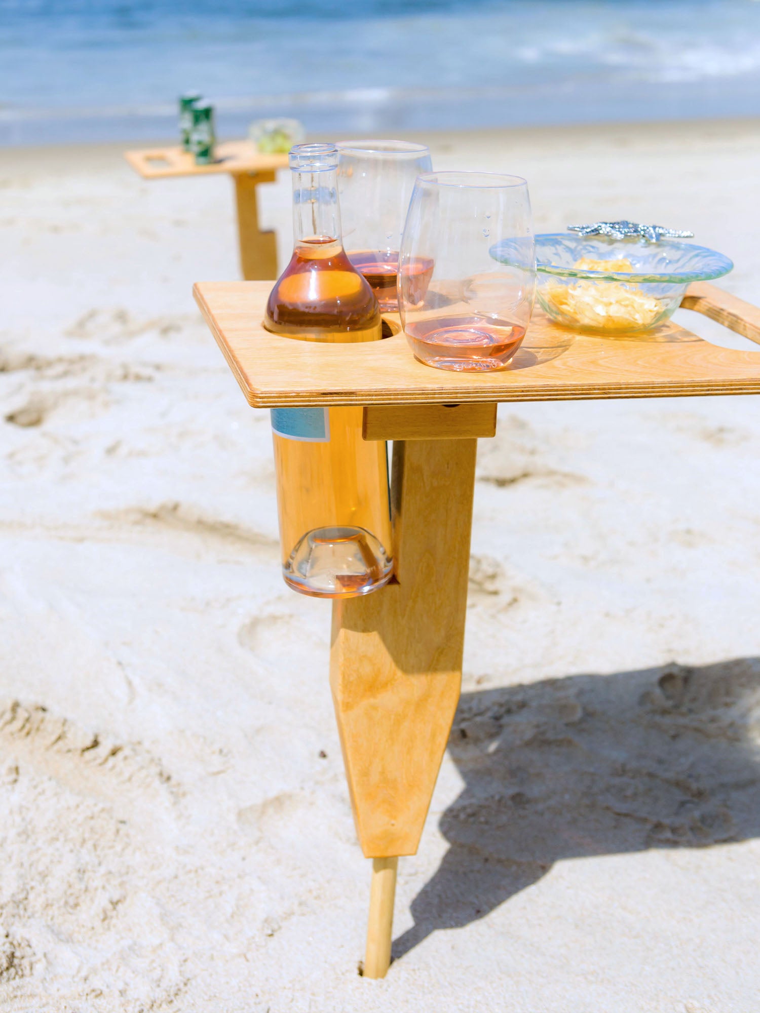 Esplanade portable beach table in naturally sunkissed color unfolded standing up in sand at beach with refreshment drinks and food